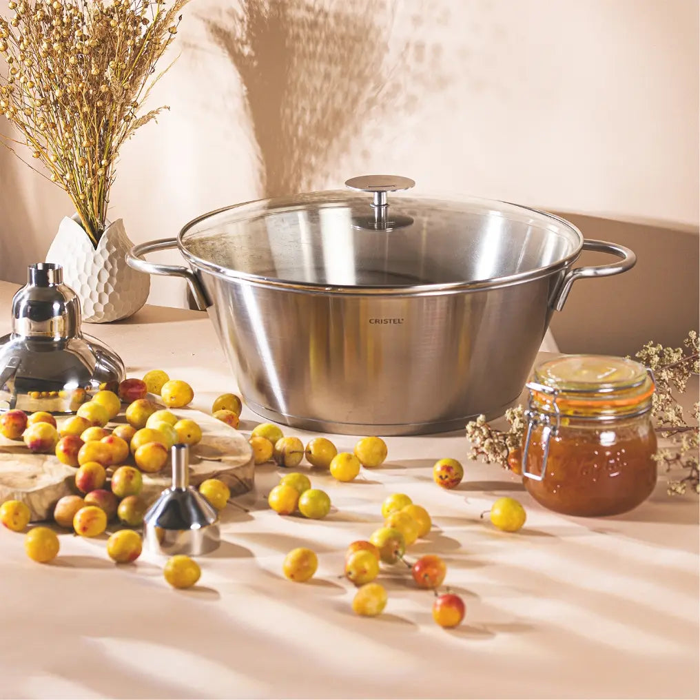 Brushed stainless steel preserve pan with glass lid beside amber jam and yellow plums on beige surface