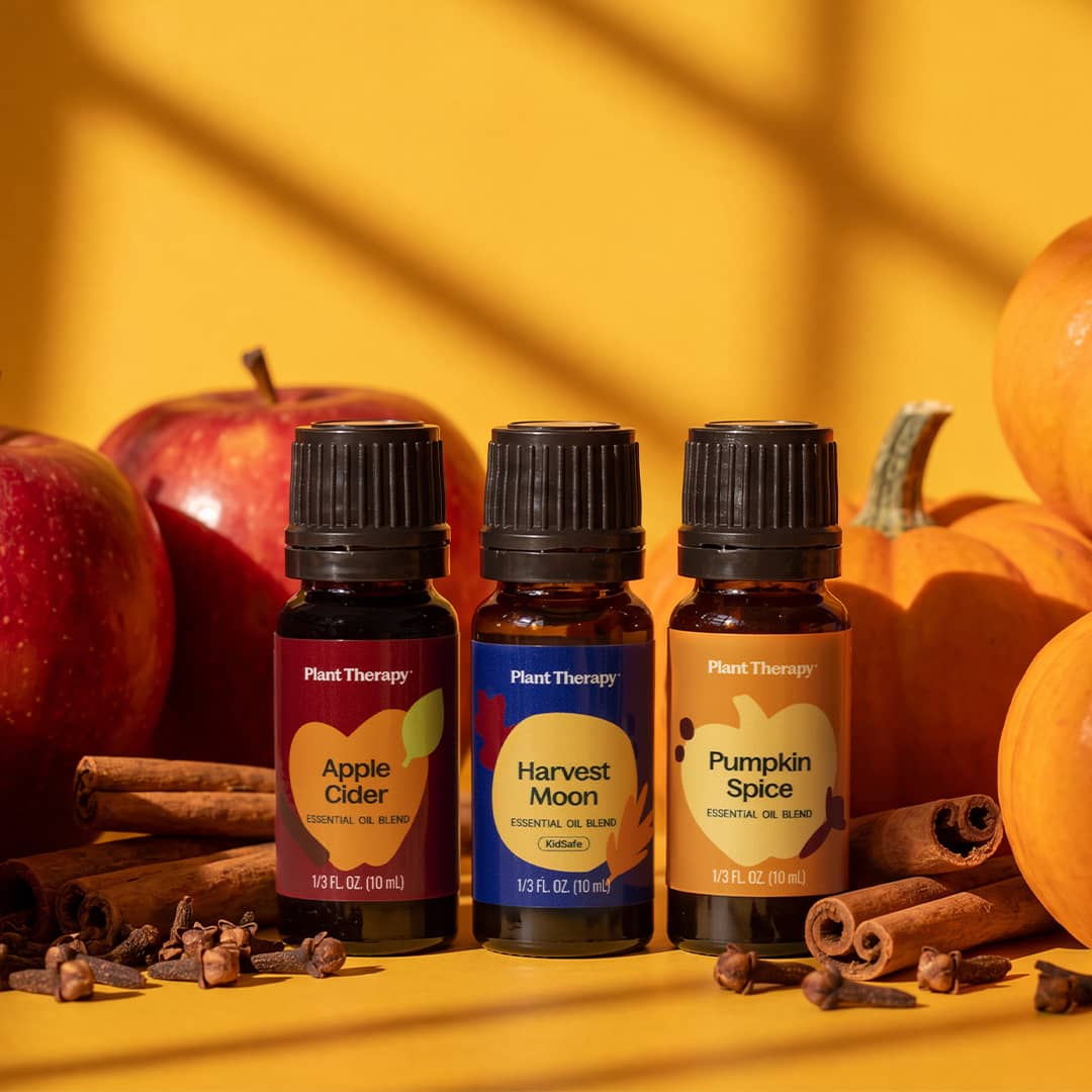 Three labeled essential oil bottles arranged on a yellow surface with red apples, pumpkins, and cinnamon sticks under warm