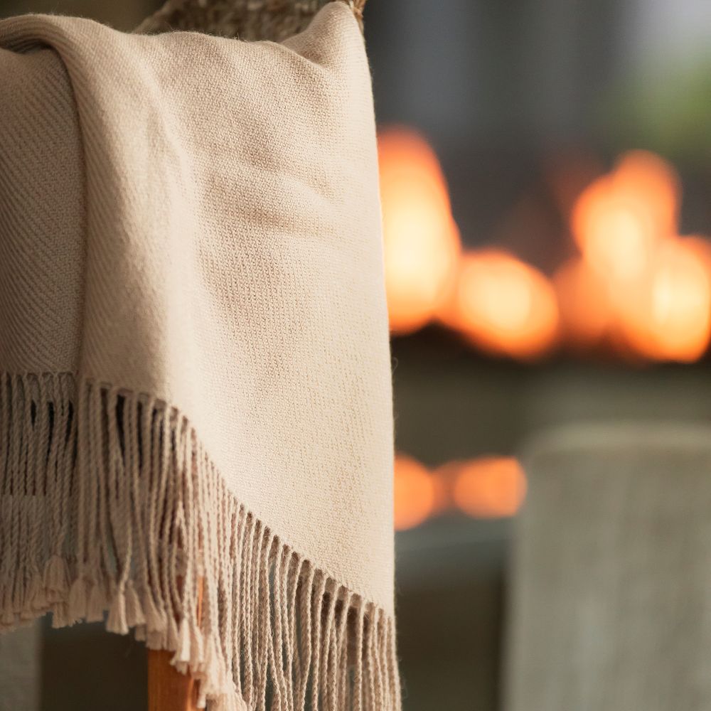 Beige undyed baby alpaca throw draped over a wooden chair with long hand-tied fringe against warm bokeh lights