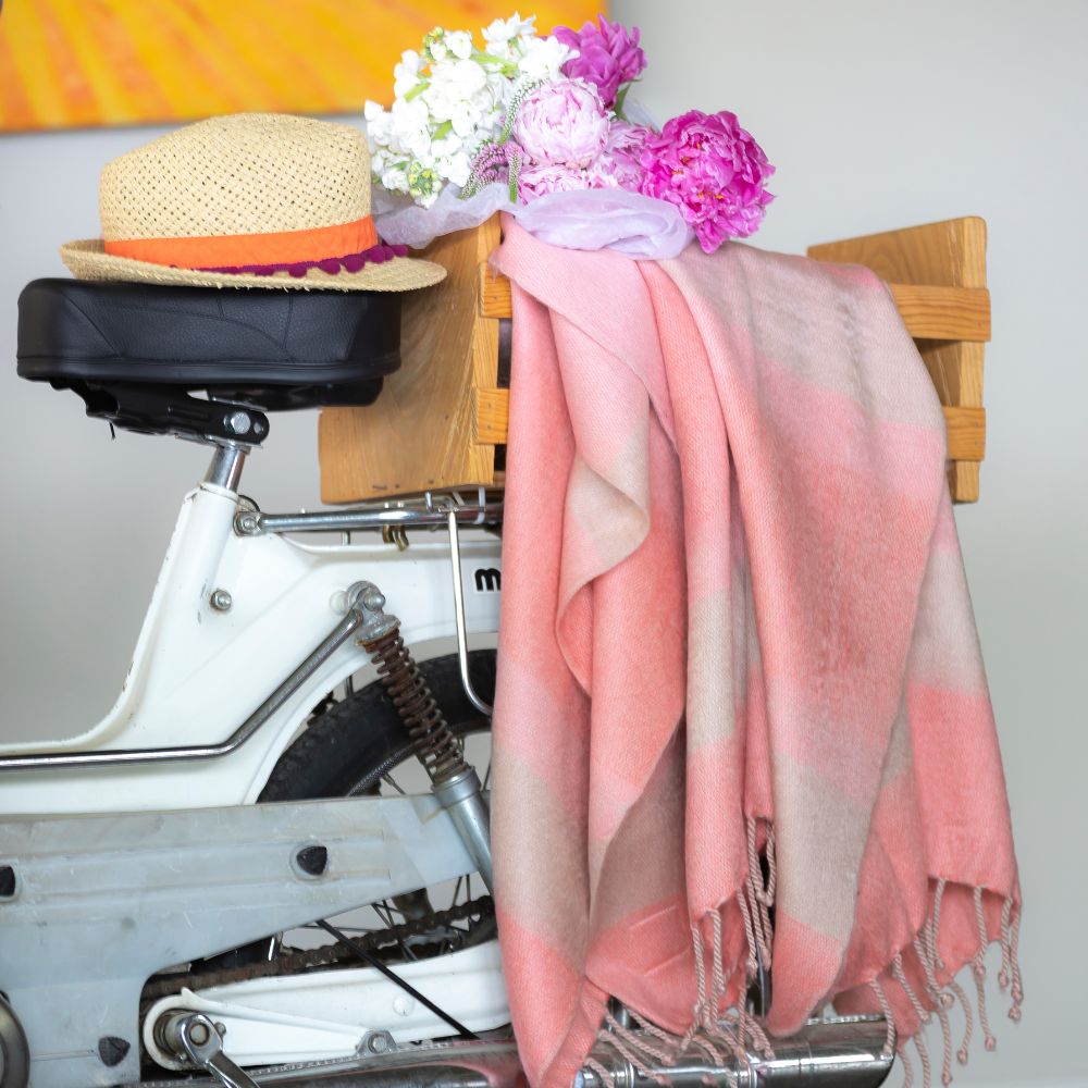 Faded coral and beige striped alpaca throw draped over a white vintage moped crate with straw hat and pink peonies