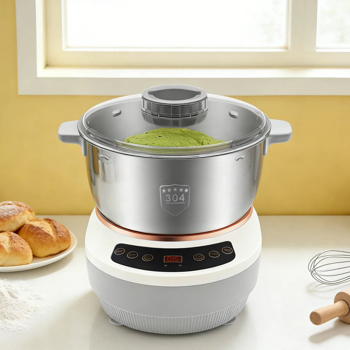 Silver electric dough kneader with stainless steel bowl holding green matcha dough under a clear lid, next to baked bread
