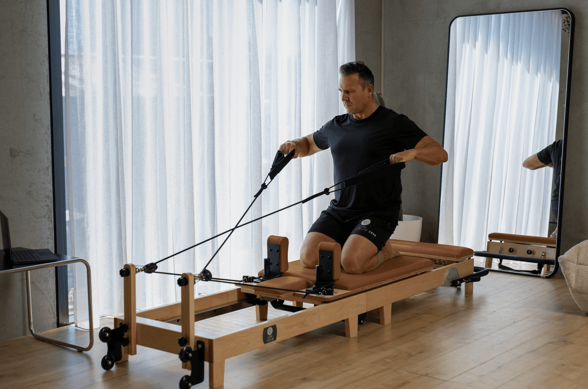 Man using tan upholstered reformer positioned by window with white drapes highlighting color options