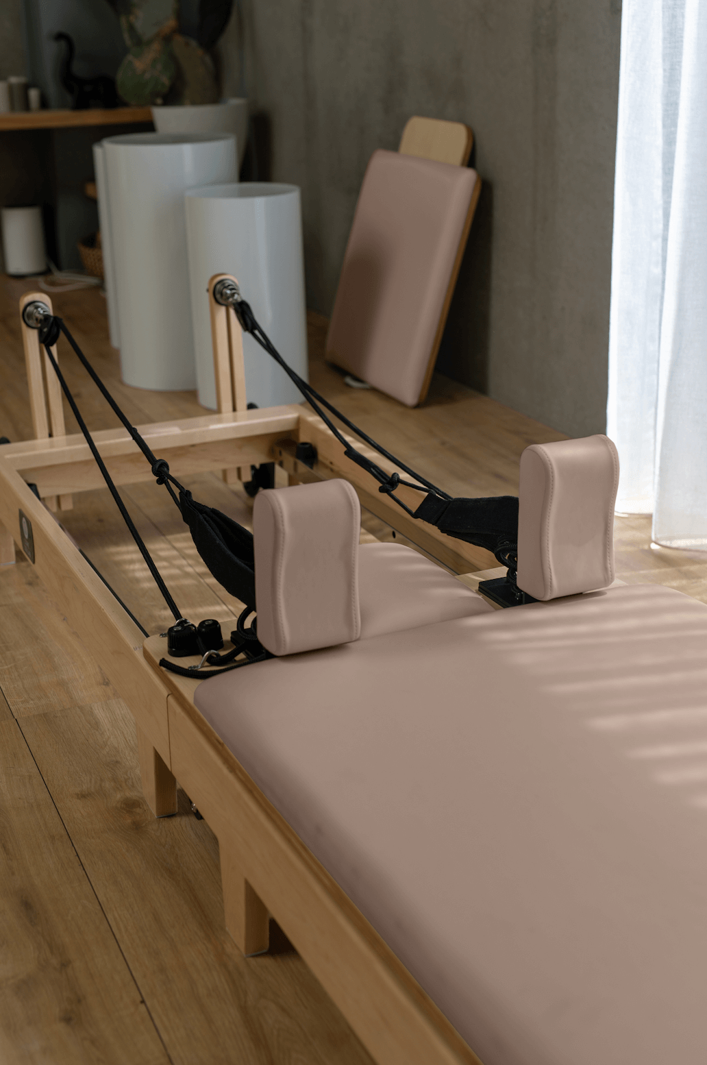 Close-up of light pink padded reformer with natural oak frame, black straps, and concrete studio background
