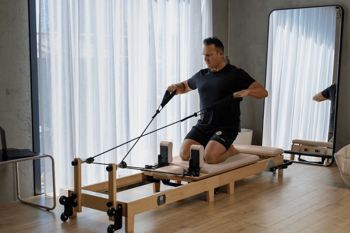 Male instructor in black wear kneeling on pink reformer pulling handles near mirror and sheer curtains