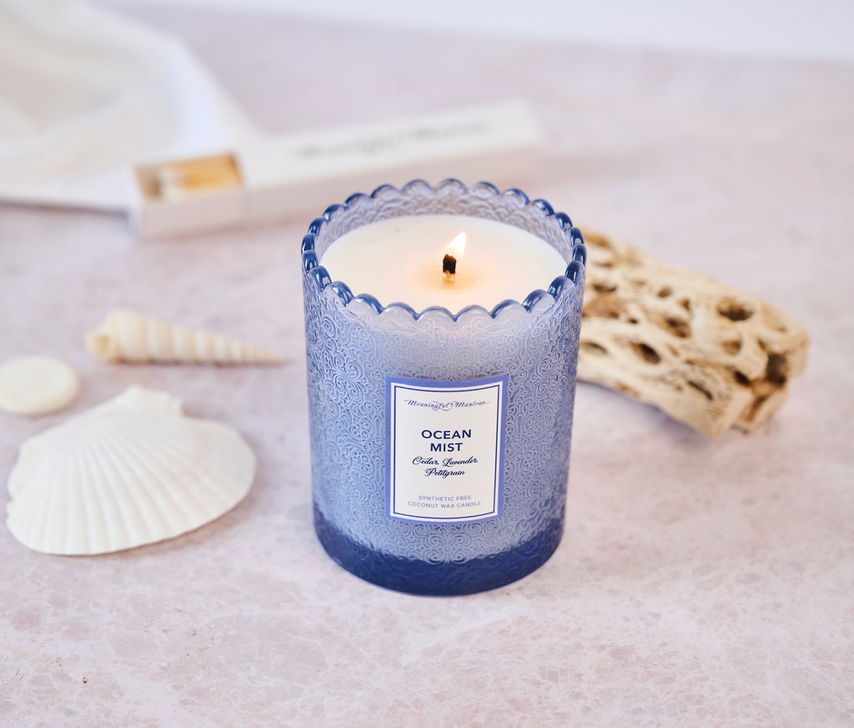 Lit coconut wax candle on pink marble surrounded by white seashells and driftwood