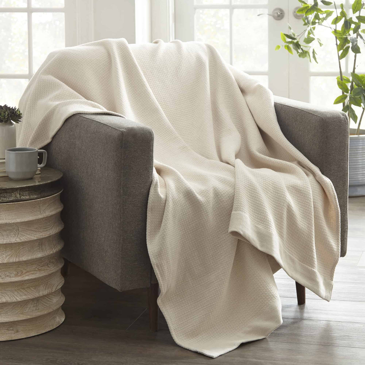 Cream woven blanket draped over grey armchair next to wooden table and mug in living room