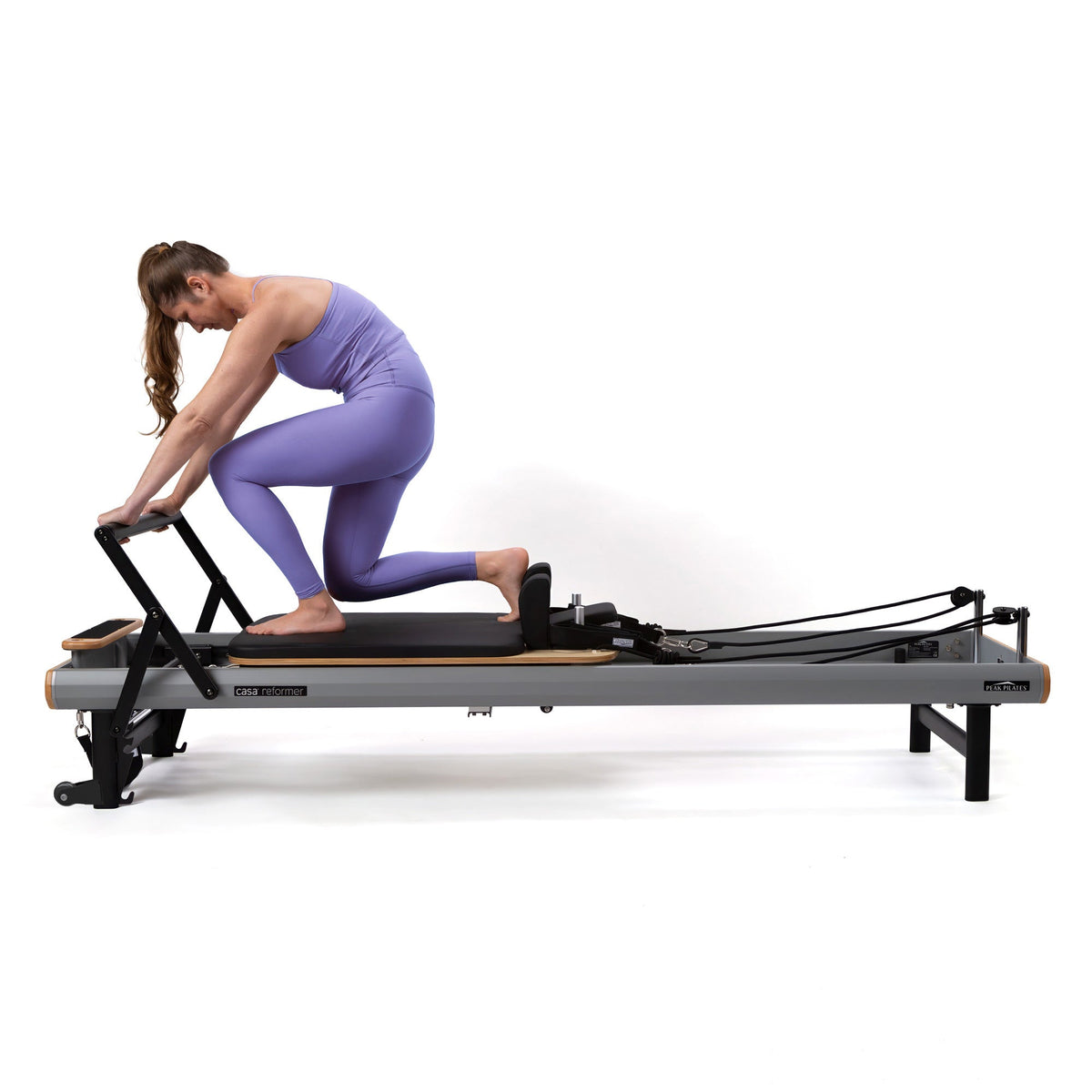 Side profile of woman in purple activewear kneeling on sliding carriage of grey and wood reformer against white background