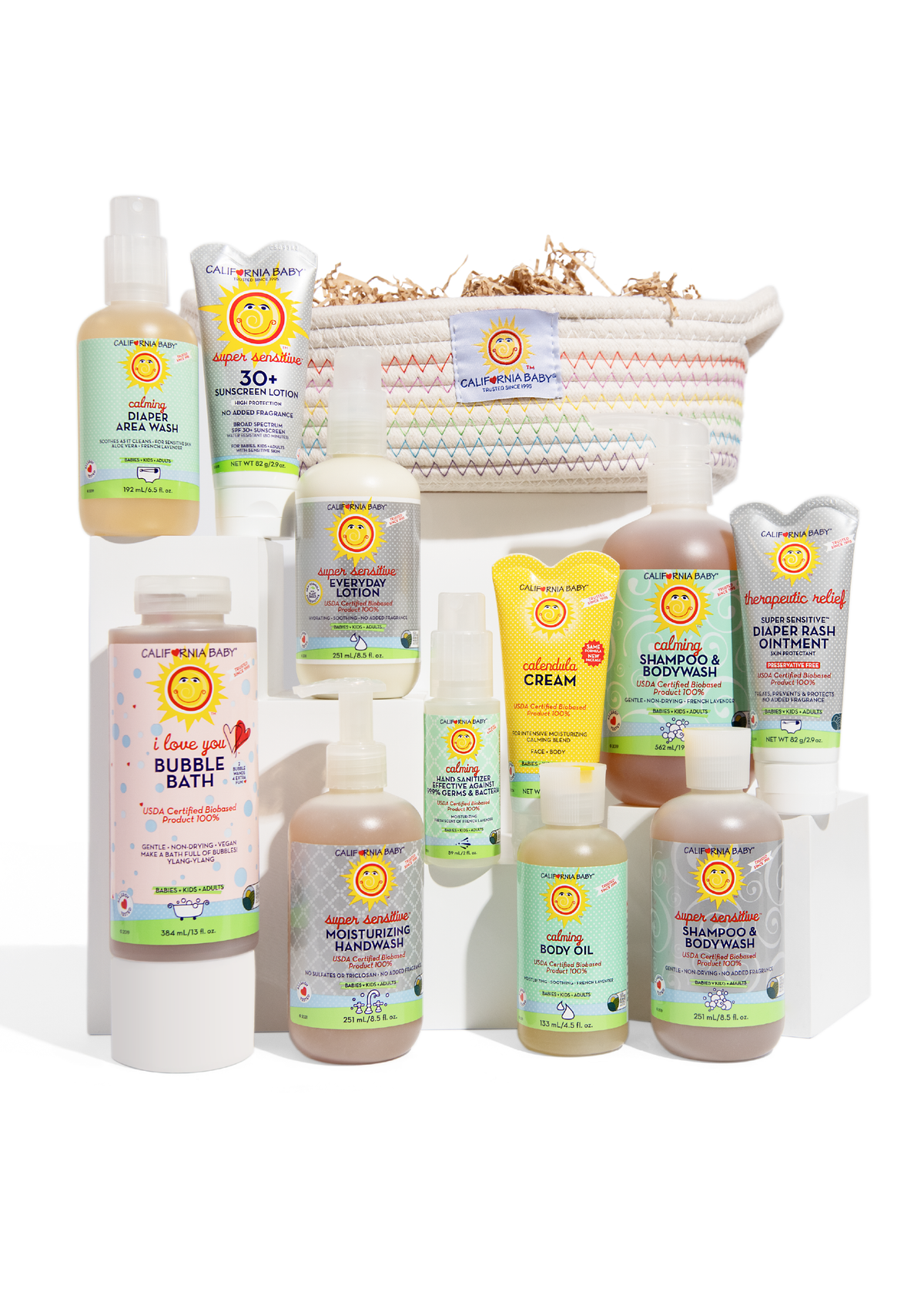 Assorted baby care bottles and tubes arranged on white blocks in front of a rainbow-stitched gift basket