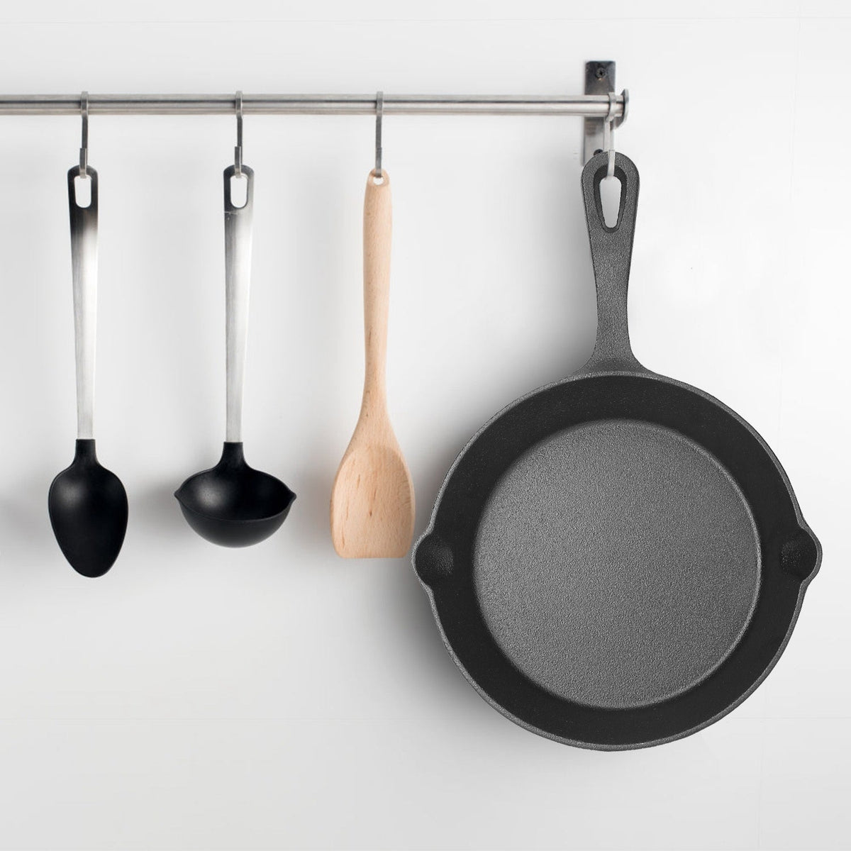 Matte black cast iron skillet hanging by looped handle on stainless steel rail next to silicone spoons and spatula