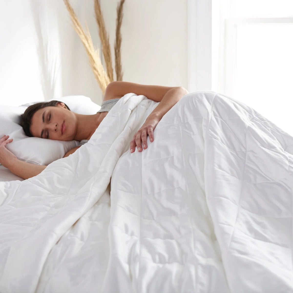 Woman sleeping peacefully under a white duvet comforter with her hand resting on the fabric in a serene high-key bedroom