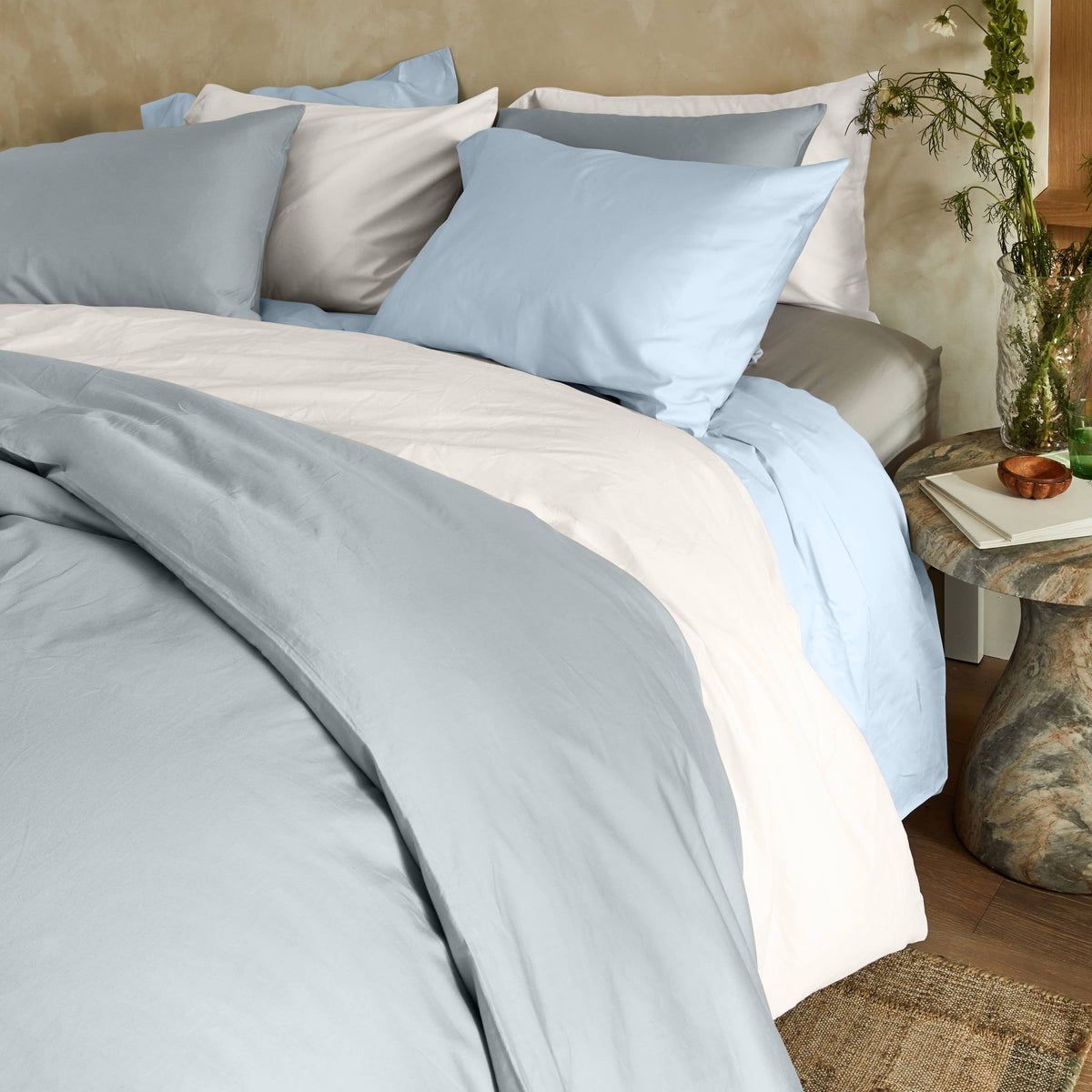 Multi-tone blue layered bedding set positioned next to a rustic stone side table with greenery