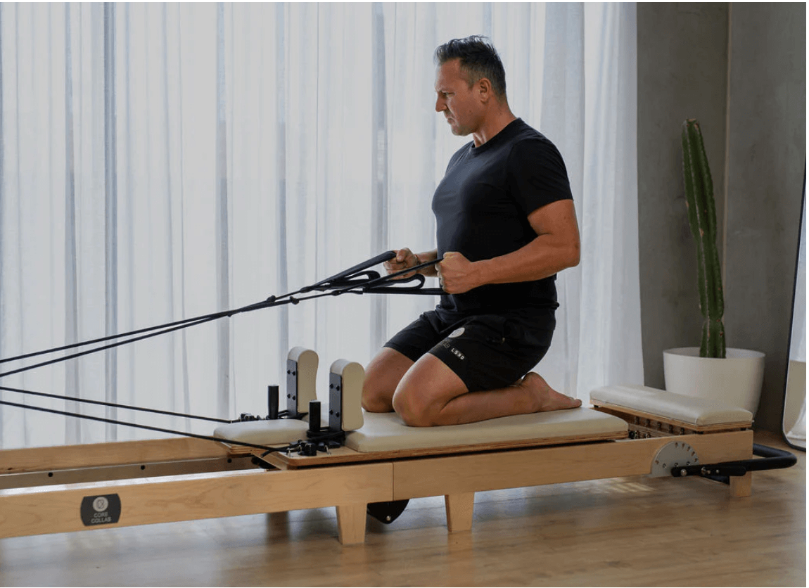 Man kneeling on beige carriage pulling black handles towards chest to demonstrate upper body resistance training on reformer