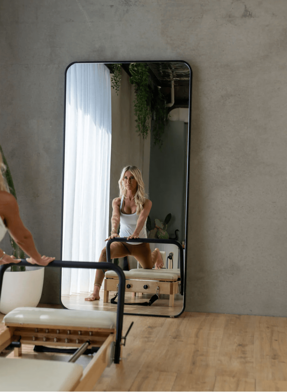 Woman using black metal footbar for support while stretching, viewed through reflection of large floor mirror with greenery
