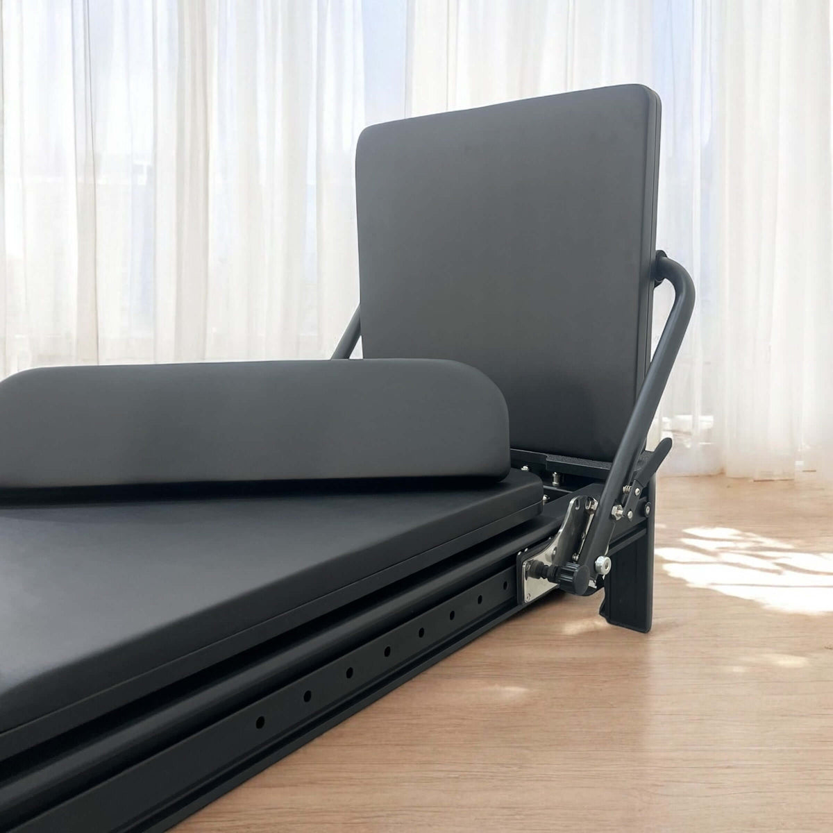 Side angle of black reformer headrest with padded backrest, curved metal supports, and sliding track bolts.