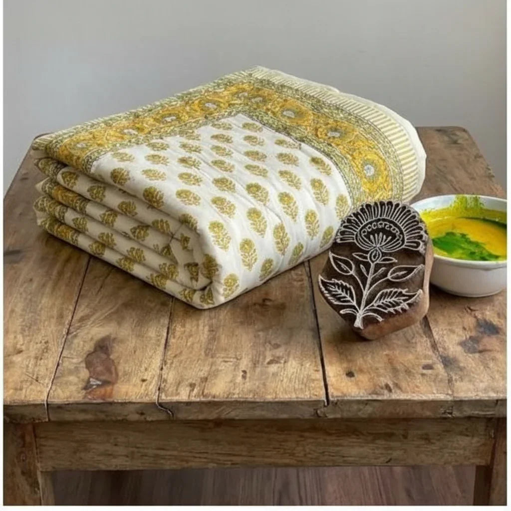 Folded comforter next to traditional carved wooden hand block and bowl of yellow-green dye on rustic table