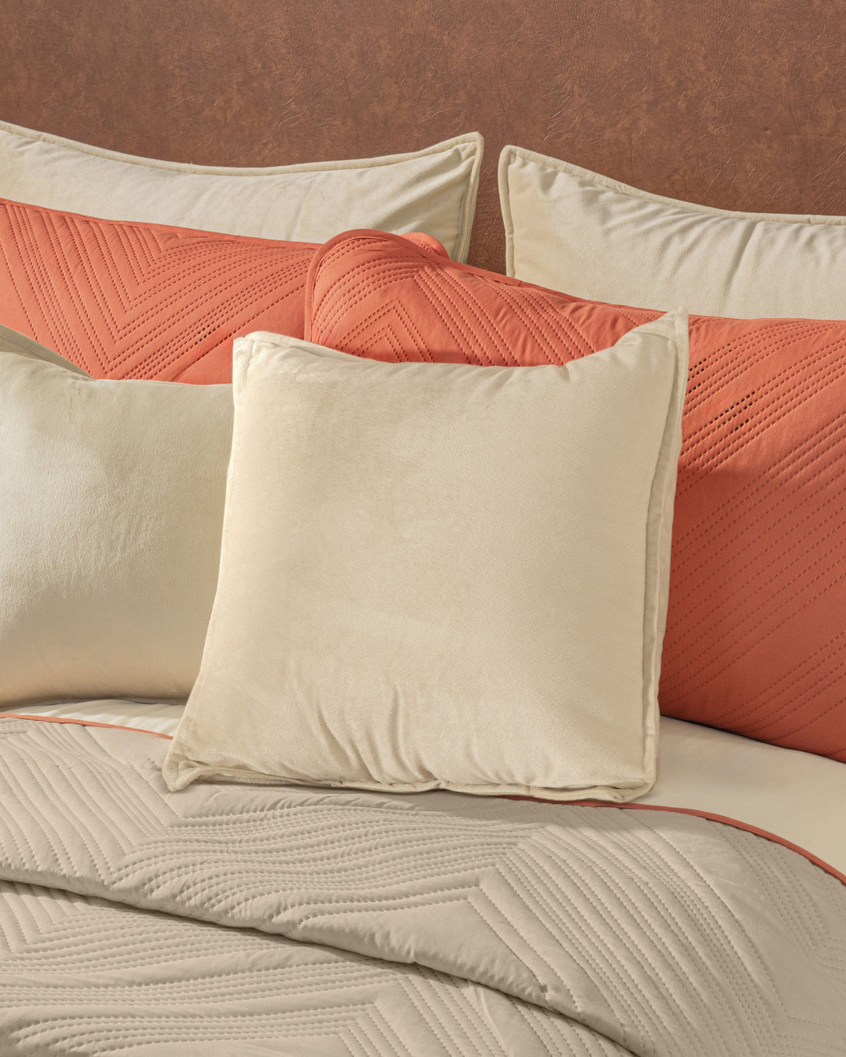 Square cream velvet pillow centered against a terracotta decorative pillow and beige bedspread with orange trim