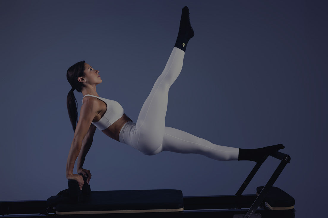 Female athlete in white activewear performing core exercise on black reformer against dark blue background