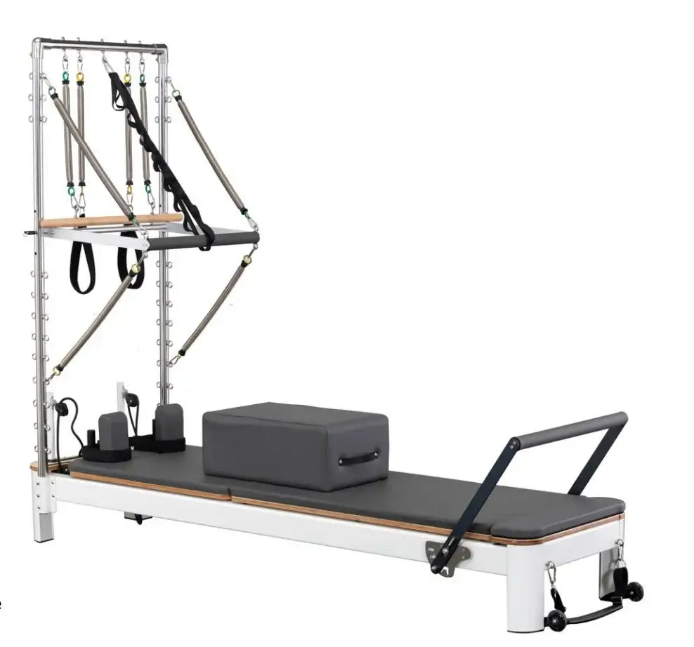 White metal frame Pilates reformer with tower, dark grey carriage, wooden base trim, and black hanging straps on white