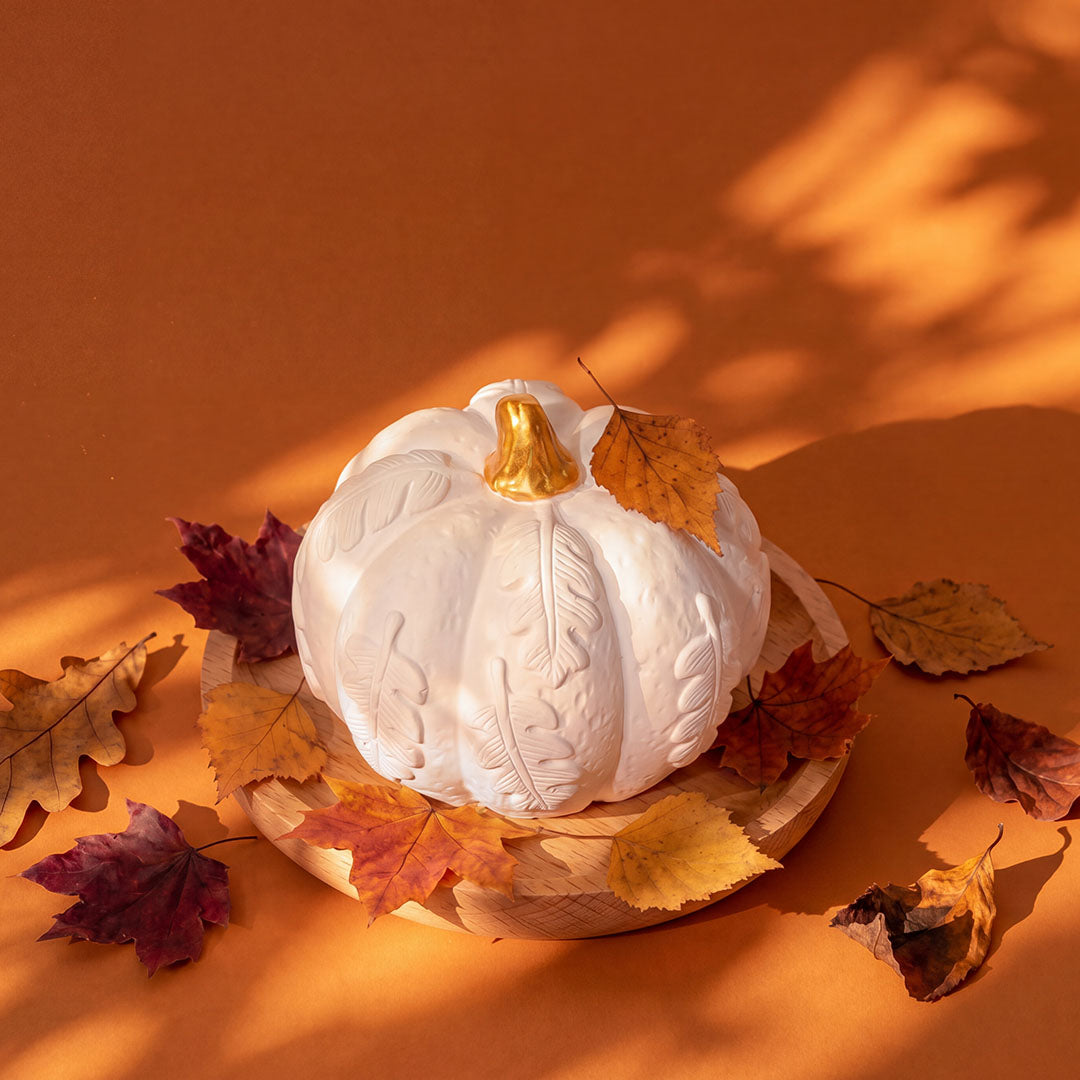 White pumpkin diffuser on wooden tray surrounded by red, yellow, and brown dried fall leaves on orange backdrop
