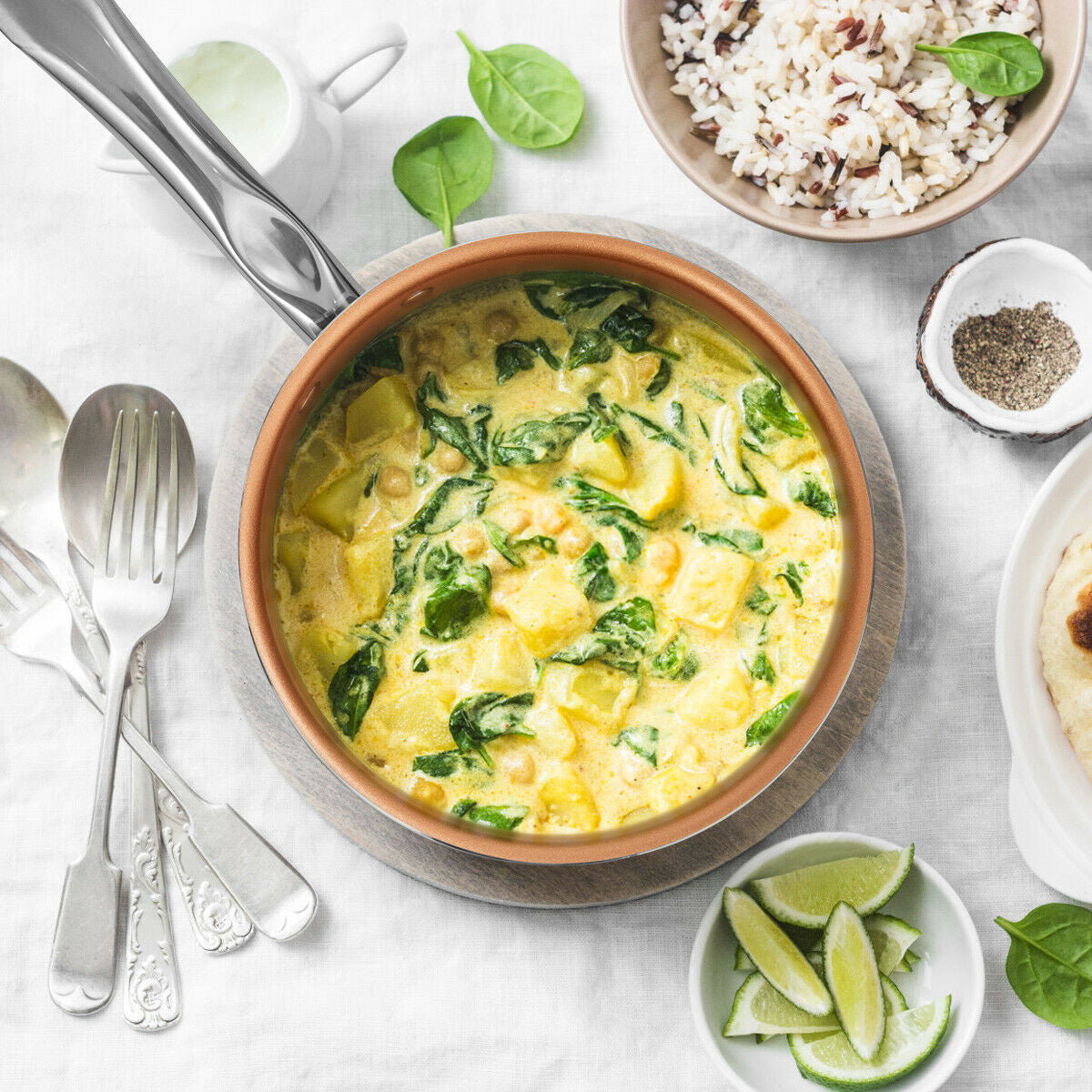 Top-down view of copper non-stick saucepan with chickpea spinach curry on grey trivet surrounded by rice, lime wedges, and