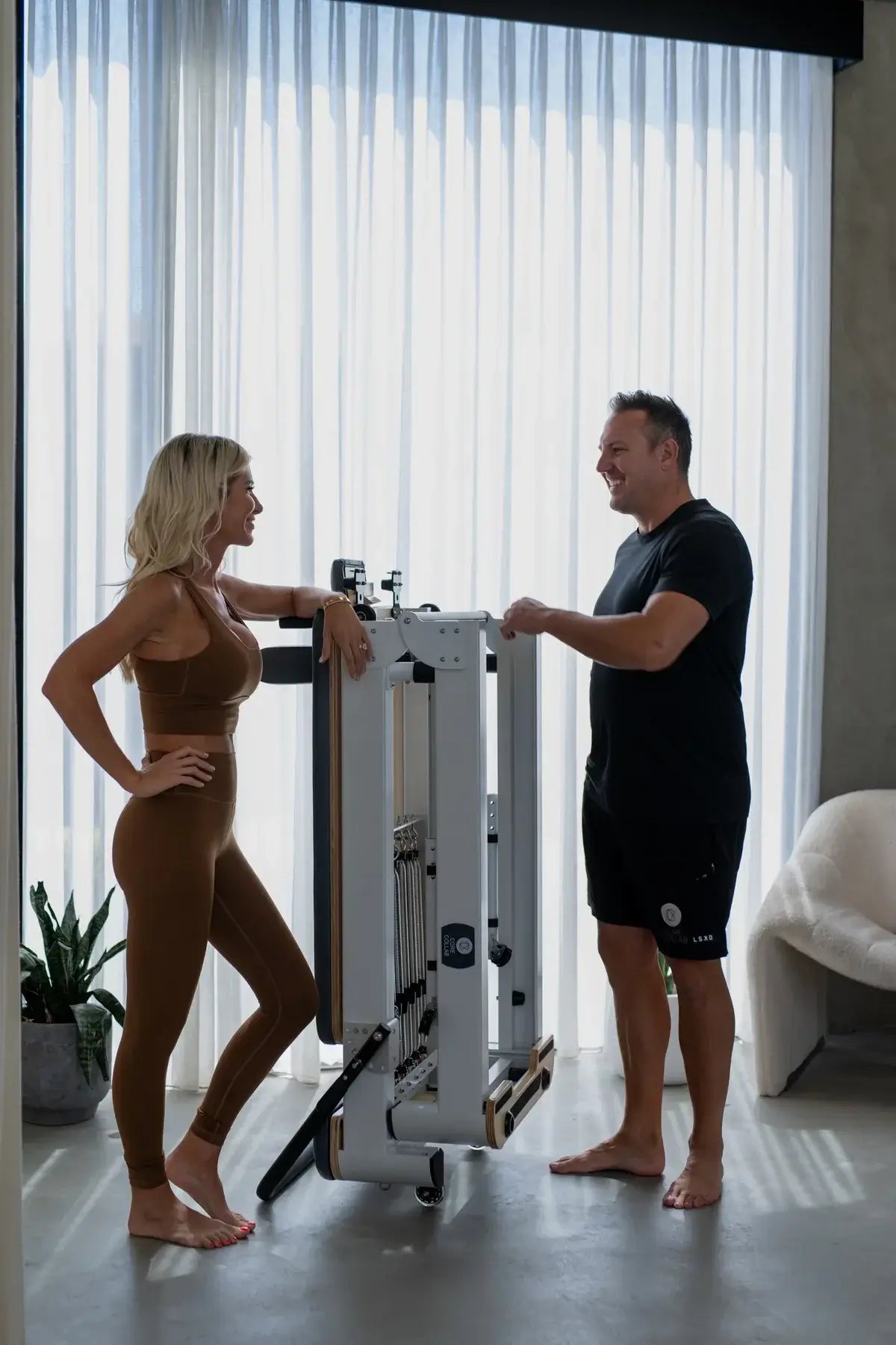 Man and woman standing beside folded white steel Pilates reformer with black upholstery against sheer curtains