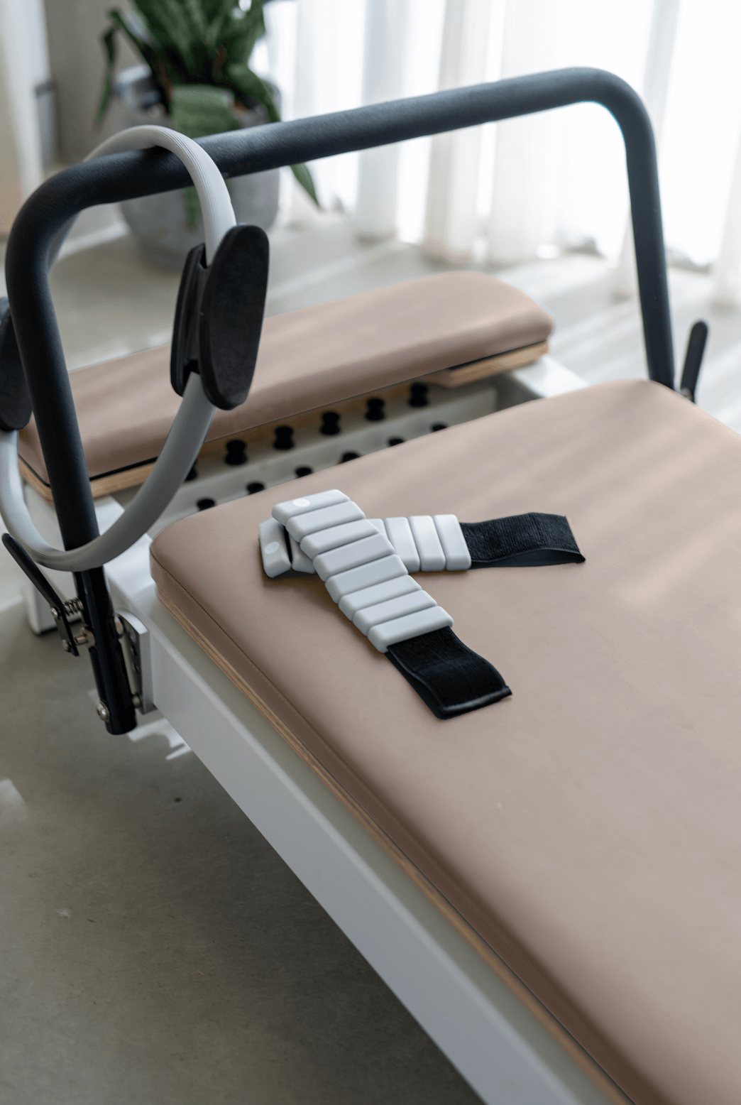 Dusty rose beige Pilates reformer carriage with white ankle strap and grey resistance band against bright window