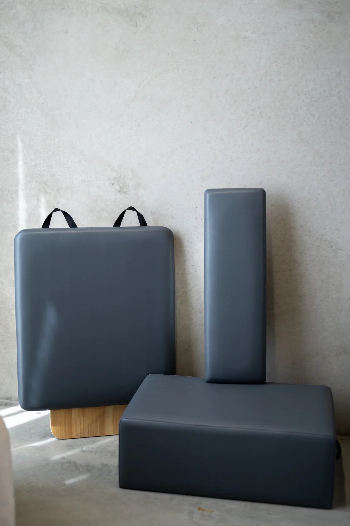 Three dark grey leather-wrapped pilates accessories including platform, block, and cushion leaning against concrete wall