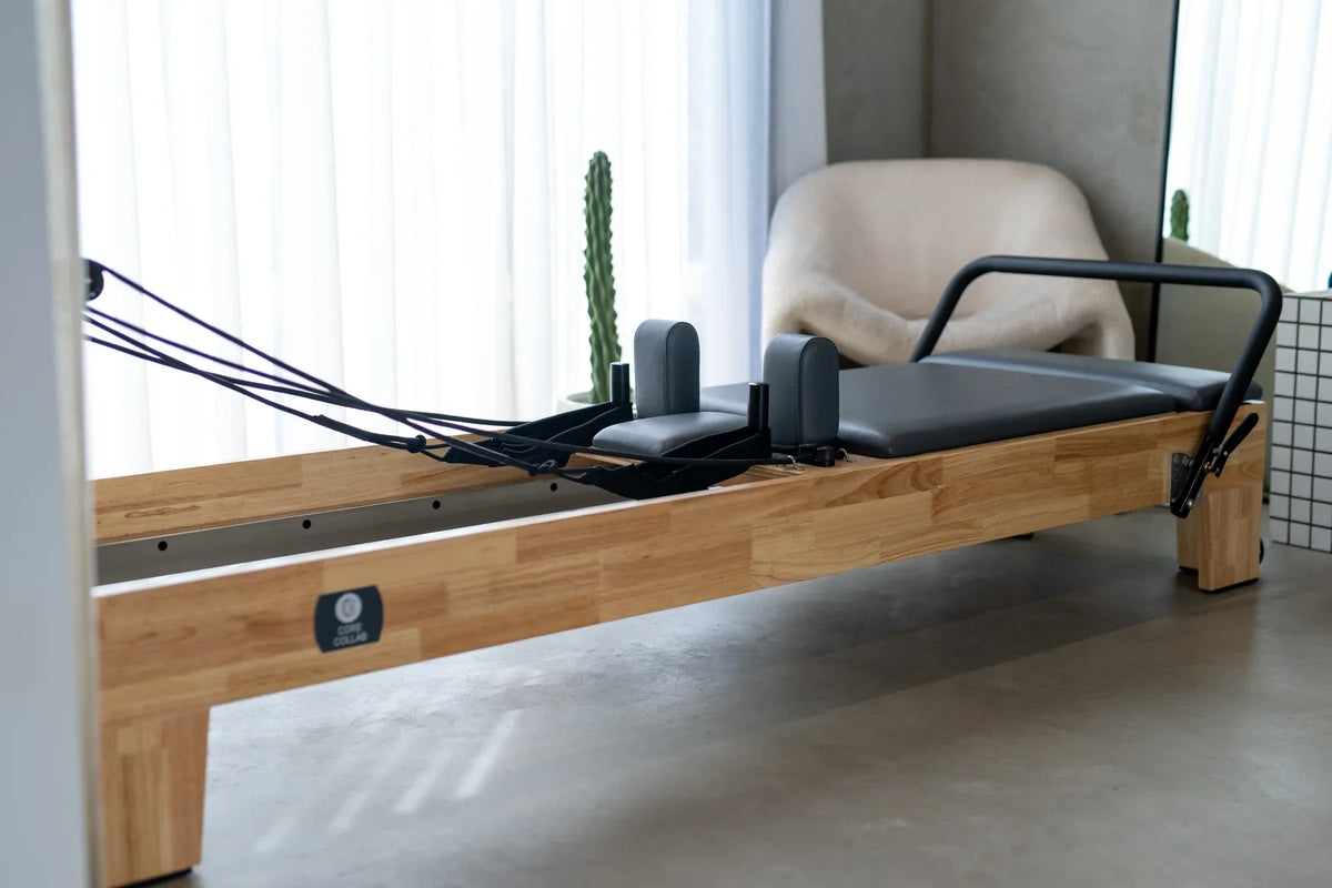 Side profile of minimalist pilates reformer showing light wood finish, black upholstery, and tension cables in studio
