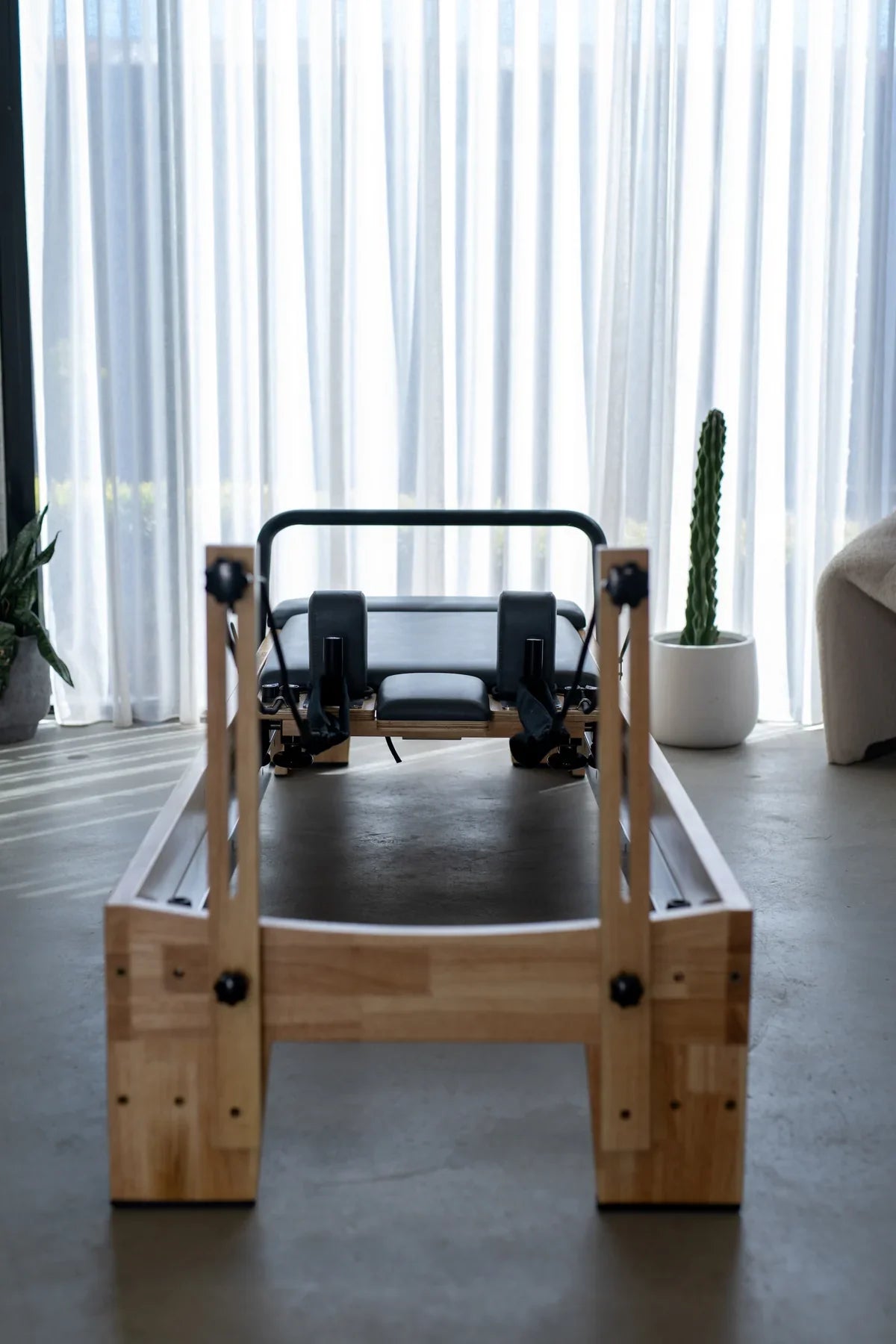 Symmetrical front view of a solid oak pilates reformer with black carriage and cable towers against white curtains