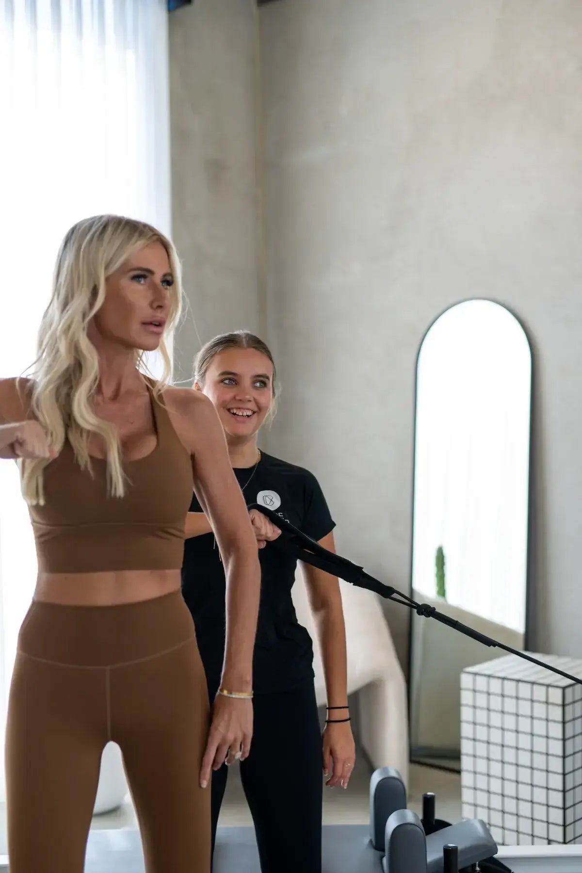 Blonde woman standing pulling black tower strap for upper body exercise while instructor adjusts equipment behind her